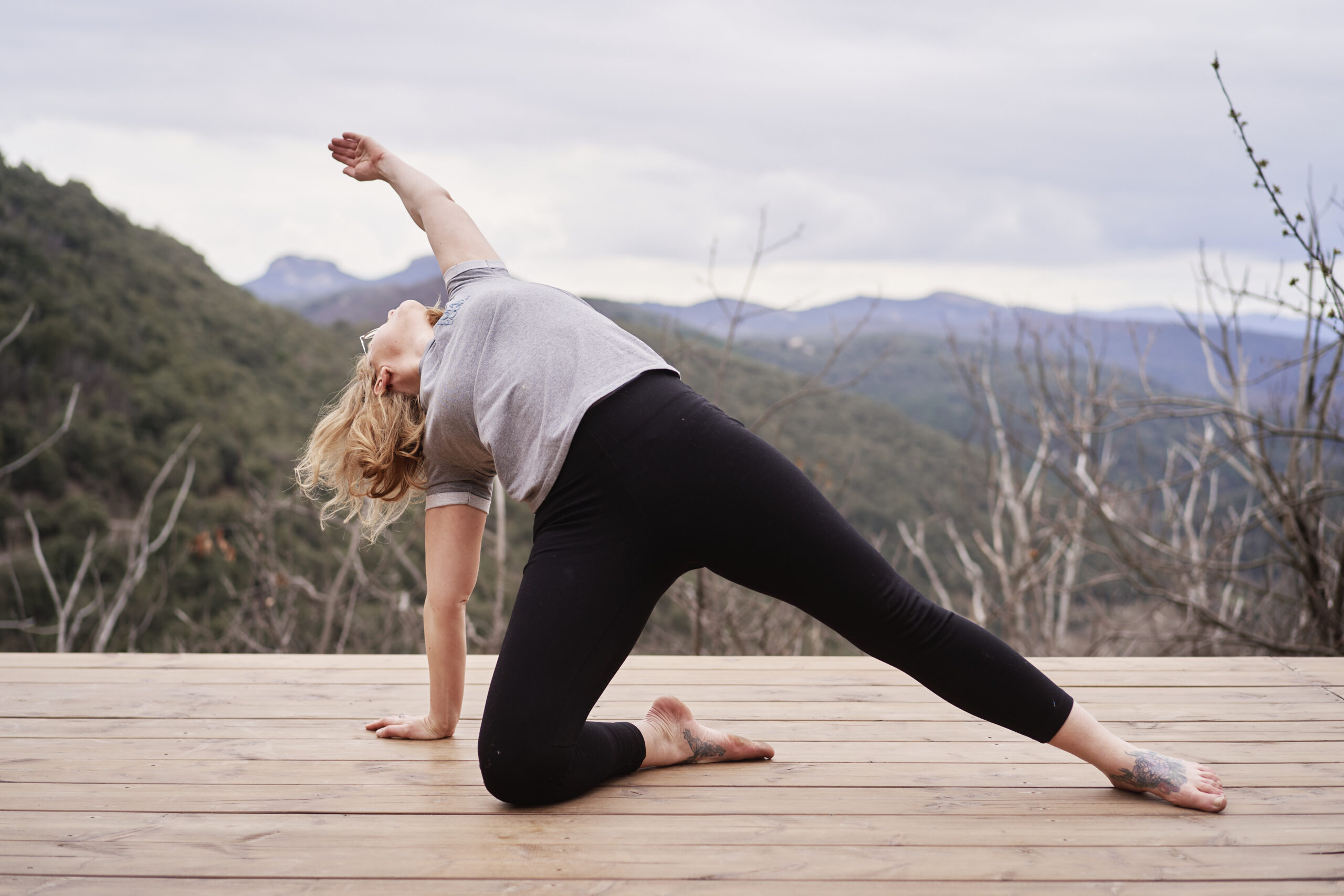 Emily, professeure de yoga à La Ciotat, effectue la posture "Camatkarasana" (Yoga Sauvage) en plein air, sur une terrasse en bois face aux montagnes.